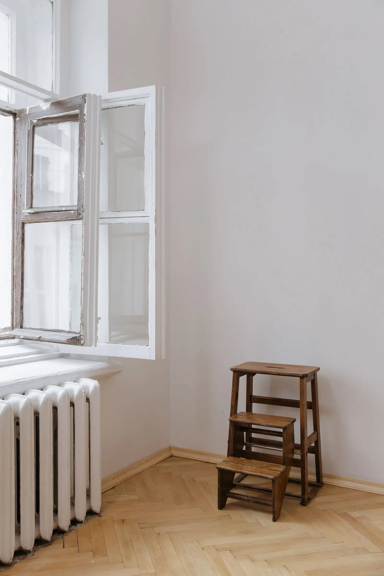 Geöffnetes Altbaufenster über einem Heizkörper, daneben ein Holzhocker in einem leeren Zimmer